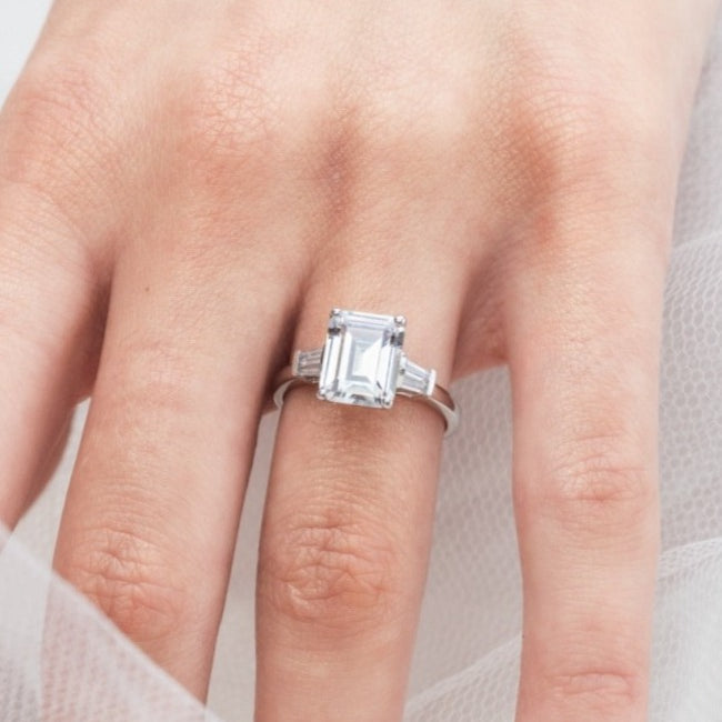 Trista - Micheli Jewellery, emerald cut trilogy ring modelled on hand. baguette side stoned trilogy ring modelled on hand. white gold emerald trilogy ring modelled on hand. trilogy ring on hand.