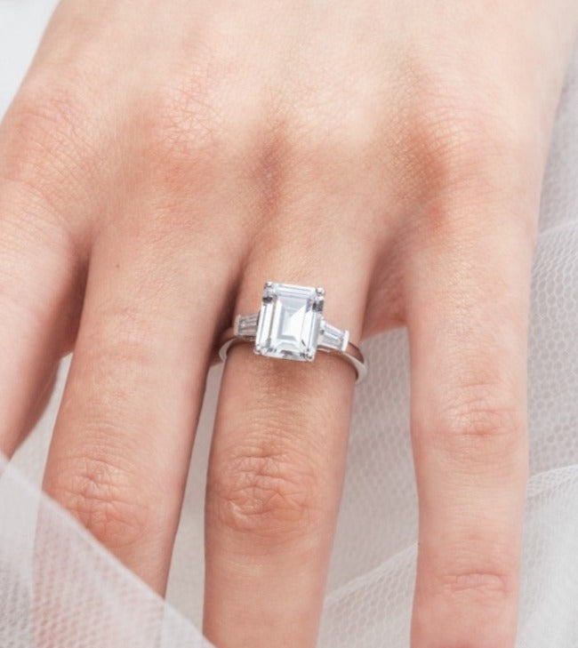Trista - Micheli Jewellery, emerald cut trilogy ring modelled on hand. baguette side stoned trilogy ring modelled on hand. white gold emerald trilogy ring modelled on hand. trilogy ring on hand.
