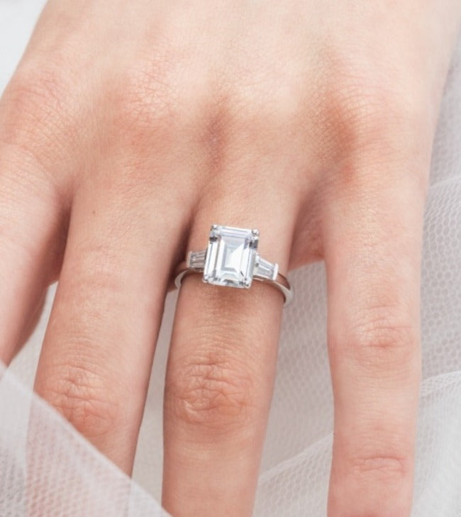 Trista - Micheli Jewellery, emerald cut trilogy ring modelled on hand. baguette side stoned trilogy ring modelled on hand. white gold emerald trilogy ring modelled on hand. trilogy ring on hand. 